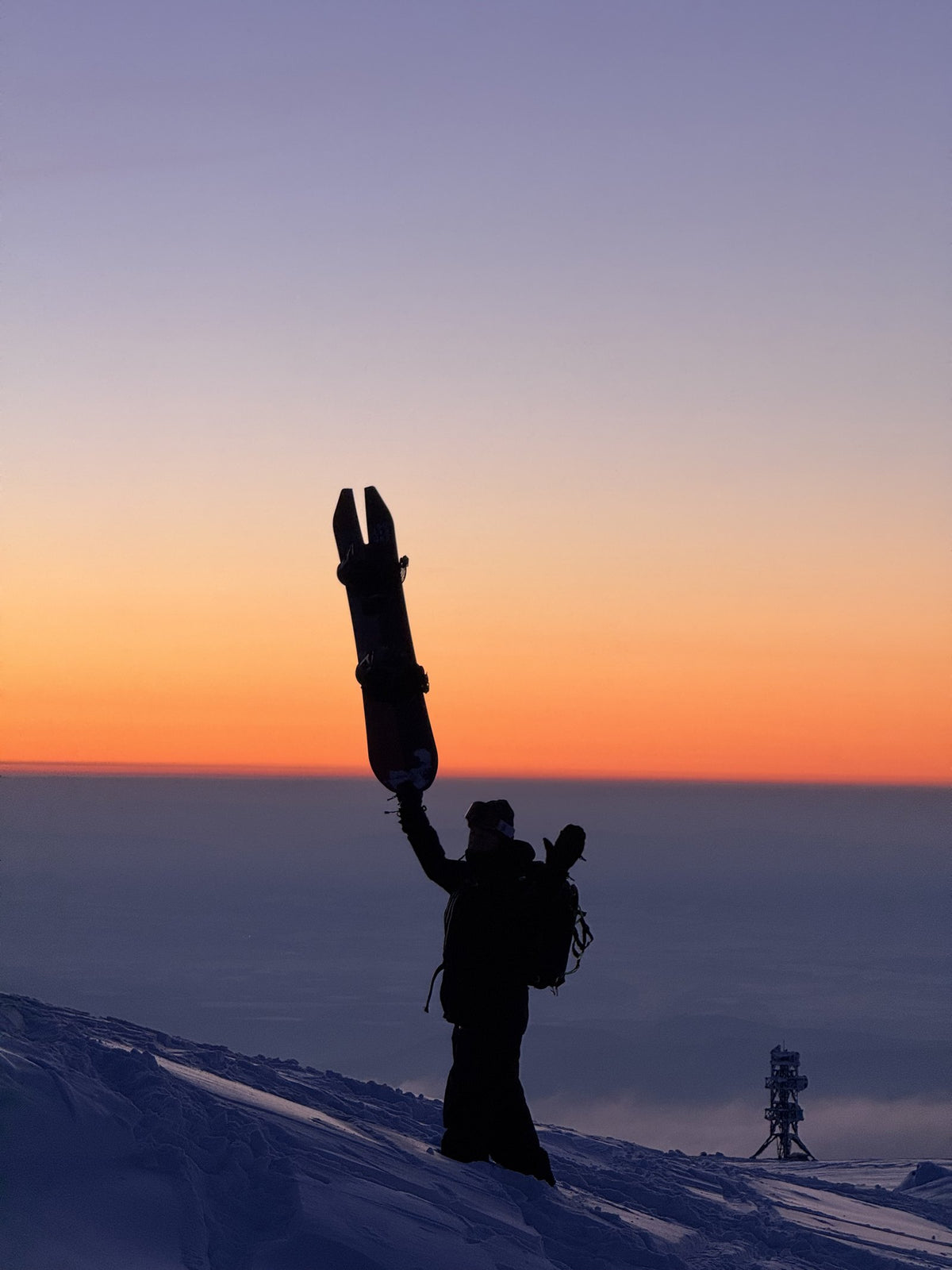 Rider holding a snowboard overhead against a sunset sky