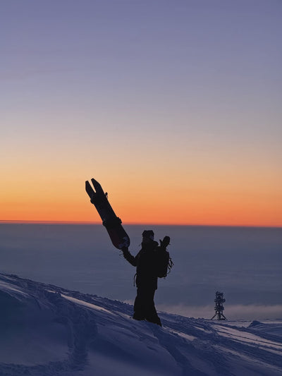 Rider holding a swallowtail Endeavor Pro snowboard overhead at sunset